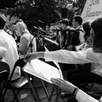 Outdoor orchestral performance with musicians playing various instruments in black and white.