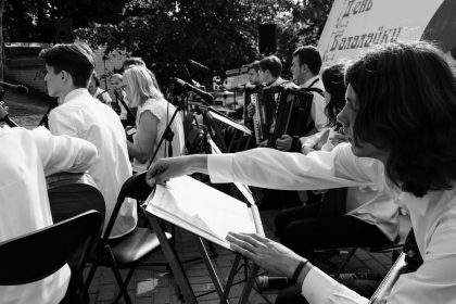 Outdoor orchestral performance with musicians playing various instruments in black and white.