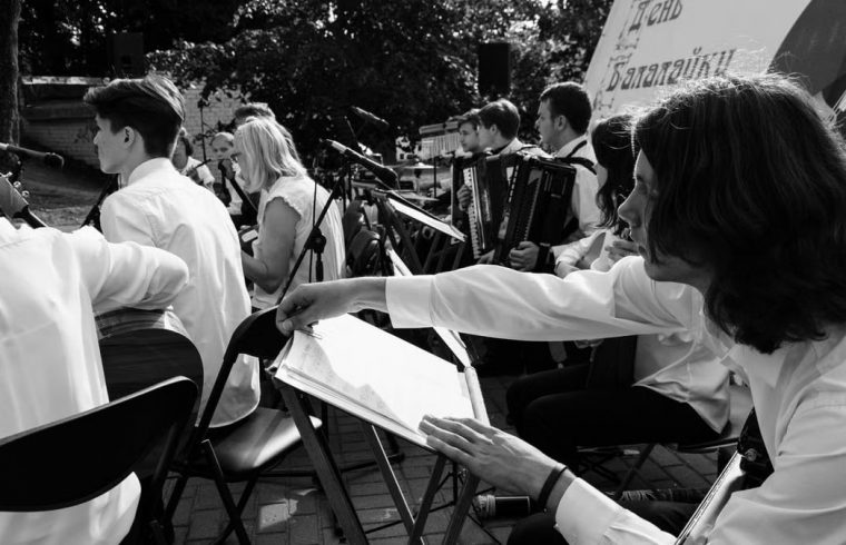Outdoor orchestral performance with musicians playing various instruments in black and white.